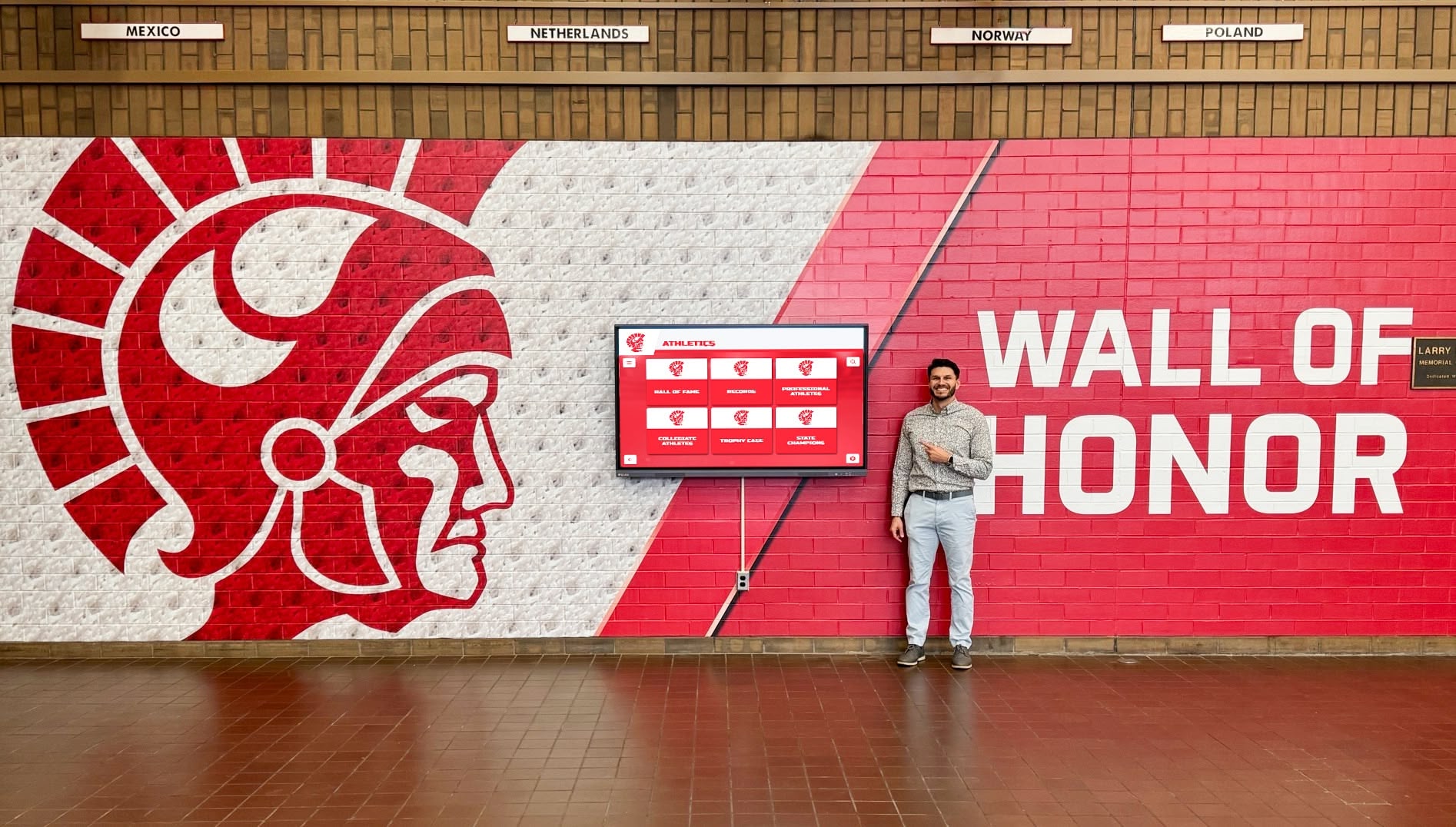 Man pointing at red Trojan Wall of Honor display in school hallway